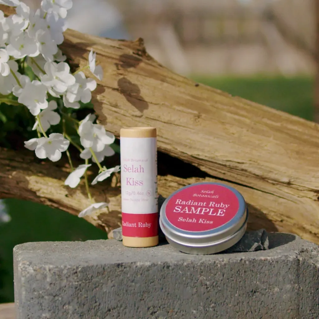 A tube of Selah Kiss lip balm and a tin labeled "Radiant Ruby Sample" are displayed on a stone surface next to white flowers and a tree branch.