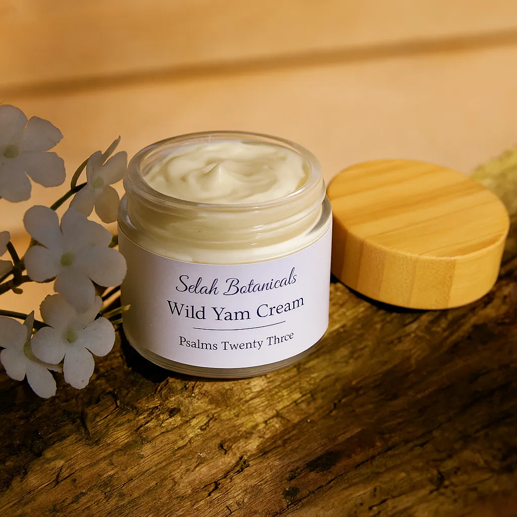 A jar of Selah Botanicals Wild Yam Cream with a wooden lid sits on a piece of wood next to white artificial flowers.
